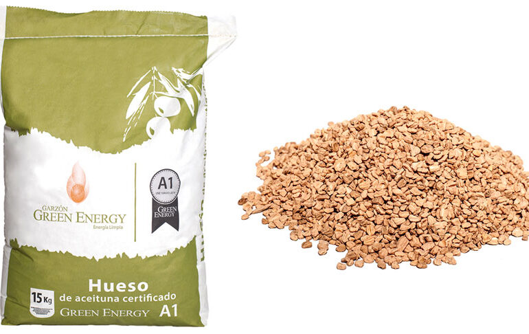 Sacchetto da 15 kg di nocciolo di oliva certificato Green Energy A1 accanto a una pila di nocciolo di oliva naturale, pronto per l'uso come biocombustibile per caldaie e stufe a biomassa.